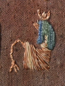Close up of a small seated figure embroidered on a rough brown fabric in varied yellow-brown thread. It has a turquoise-green wig in a rather interesting nobbly thread.