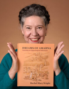 A white woman with dark hair, greying at the temples is - let's be honest - grinning at the camera. Between the flat palms of her hands she is holding up a copy of the book, "Dreams of Amarna - Stitching an Egyptological Adventure".